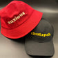 Bucket of Anxiety: Cherry red heavy cotton bucket hat with neutral coloured embroidery across the front reading ANXIOUS. Has an internal sweatband and lines sewed across the brim. Also shown is black cap with gold embroidery across the front reading CHUTZPAH. 