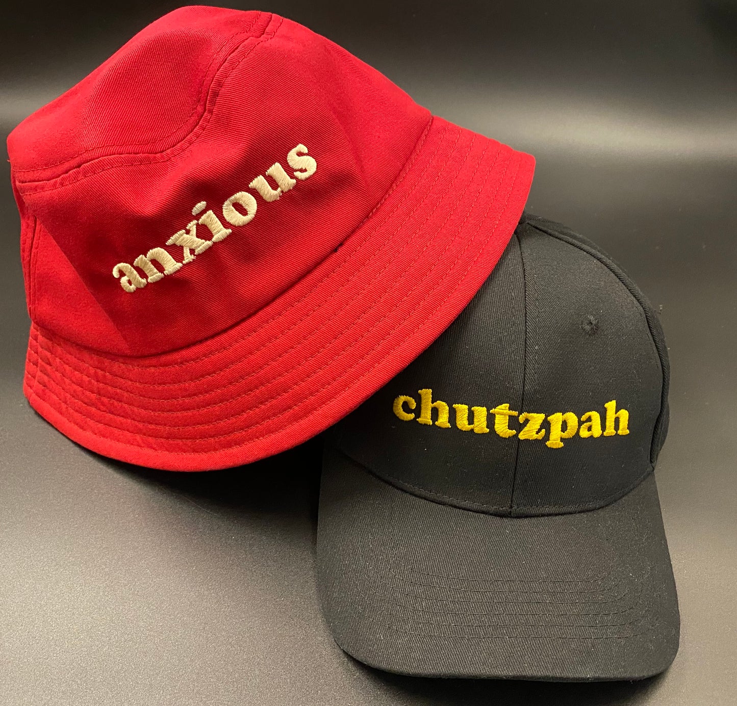 Bucket of Anxiety: Cherry red heavy cotton bucket hat with neutral coloured embroidery across the front reading ANXIOUS. Has an internal sweatband and lines sewed across the brim. Also shown is black cap with gold embroidery across the front reading CHUTZPAH. 